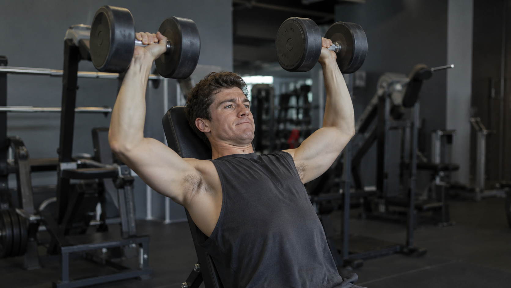 A seated lifter pressing dumbbells overhead from a gym bench